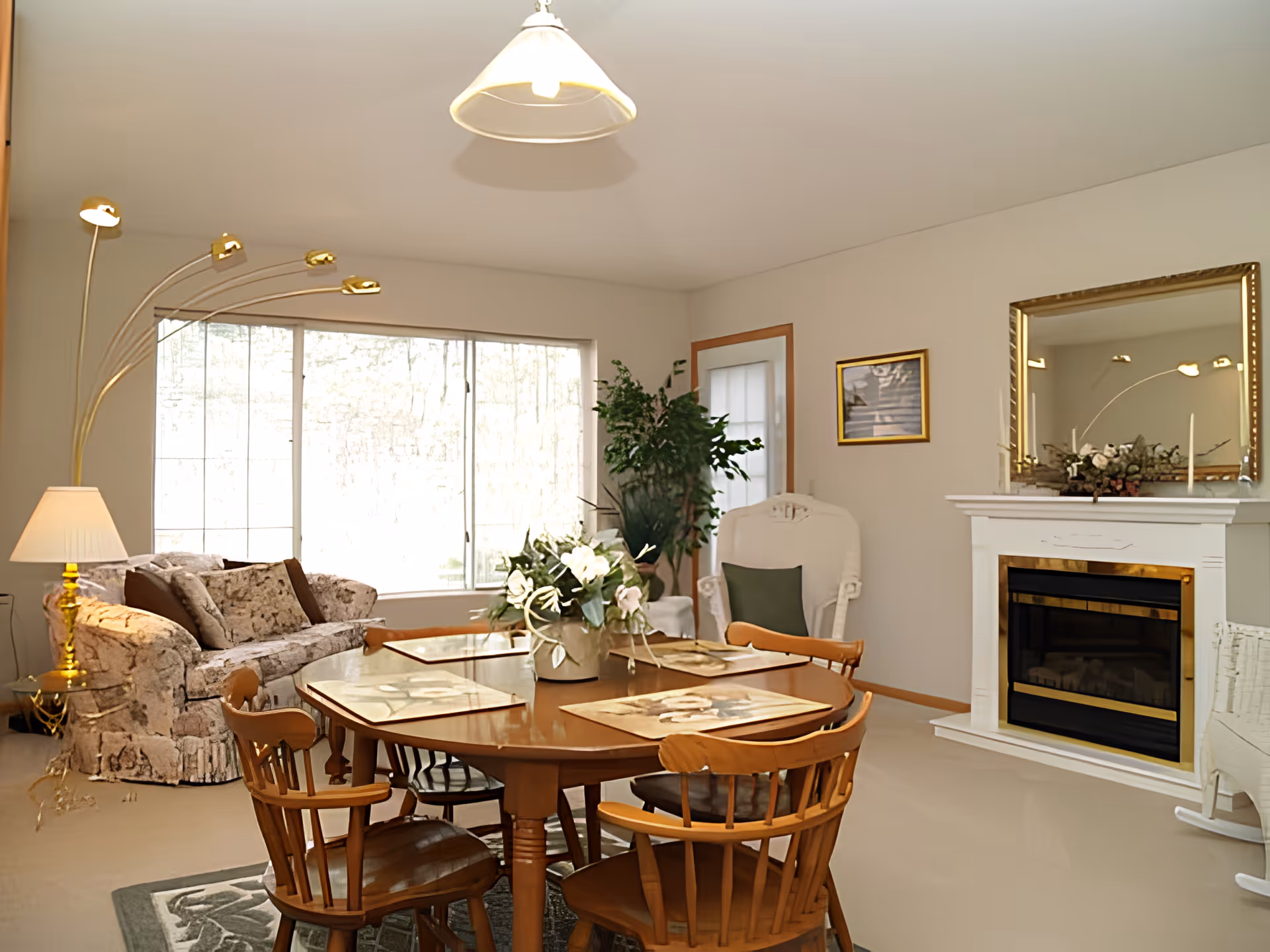 A cozy living room with a wooden dining table and four chairs in the foreground. The table has placemats and a floral centerpiece. In the background, there is a patterned sofa with cushions, a tall arc floor lamp, a white armchair with a green pillow, a large window letting in natural light, a fireplace with a gold-framed mirror above it, and a potted plant near the door.