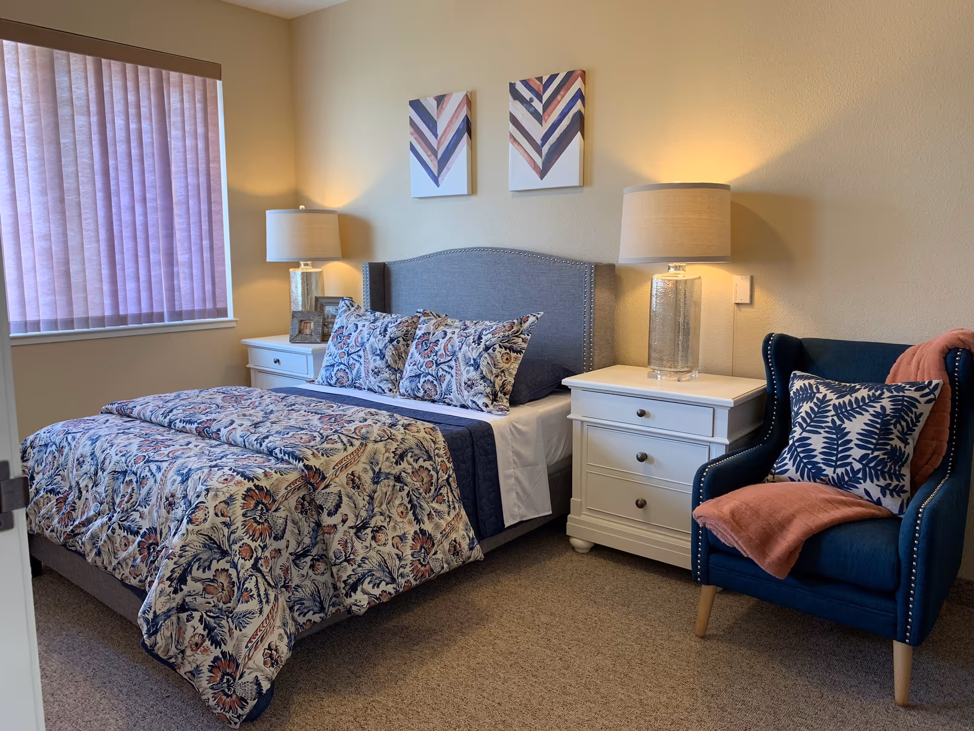 A furnished senior living bedroom with a bed, nightstands, lamps, and a blue upholstered chair.
