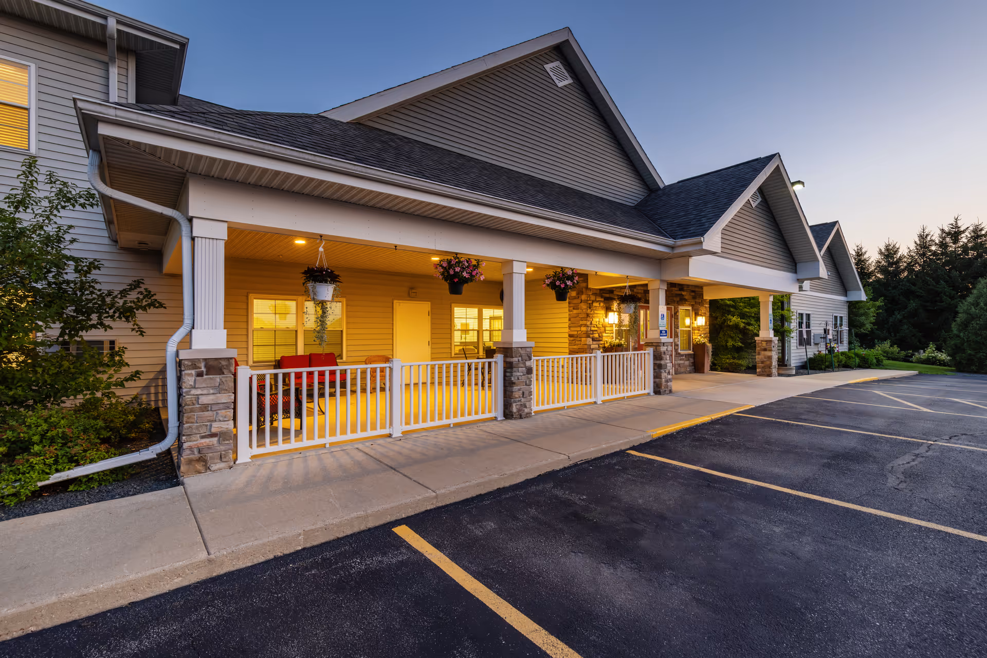 Exterior view of Milestone Senior Living Cross Plains building at dusk, showing a covered entrance with stone pillars, hanging flower baskets, and a parking area in front.