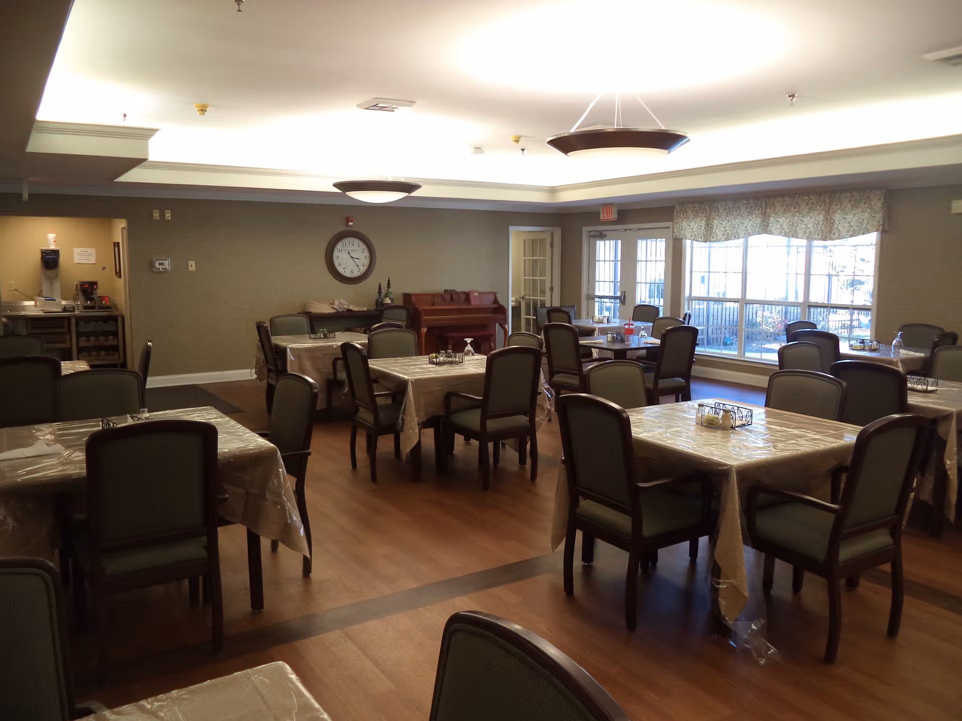 A dining room with multiple tables covered with plastic tablecloths and surrounded by chairs. There is a large window with curtains letting in natural light, a clock on the wall, a piano, and a beverage station in the corner.
