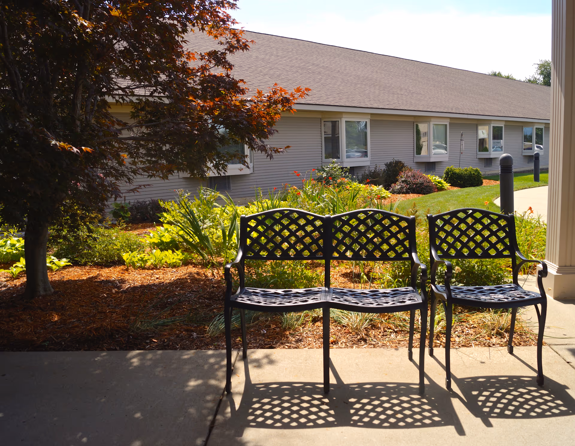 Outdoor seating area with a black metal bench and chair casting shadows on a concrete patio. Behind the seating is a landscaped garden with green plants and a tree with reddish leaves. In the background, there is a single-story building with multiple windows and a sloped roof under a clear sky.