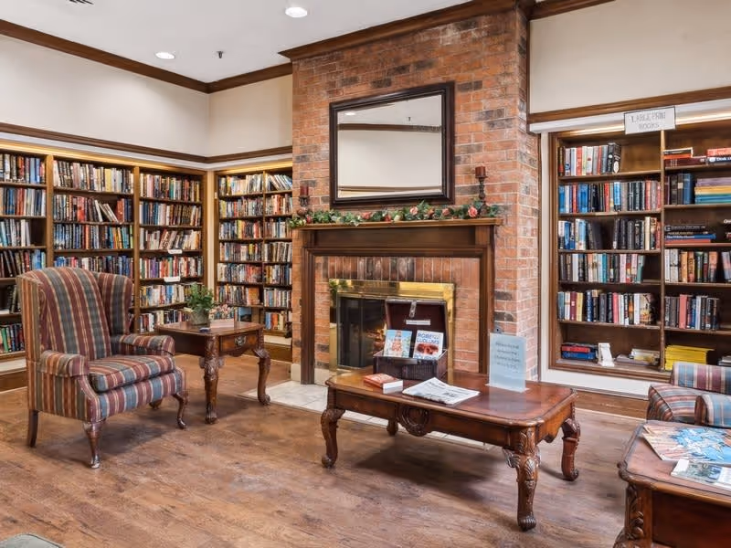 A cozy library room with wooden bookshelves filled with books lining the walls. There is a brick fireplace with a wooden mantel decorated with greenery and candles. A large mirror hangs above the fireplace. The room features wooden flooring and classic wooden furniture, including a striped upholstered armchair, a small side table with a plant, and a coffee table with magazines and newspapers.