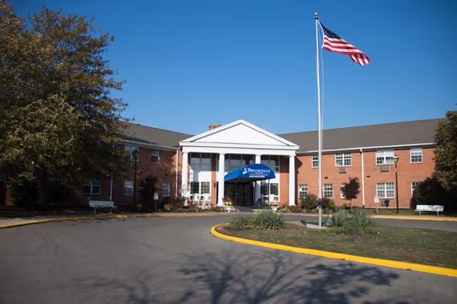 Front exterior view of a two-story brick building with white columns at the entrance and a blue awning that reads 'Brookdale'. An American flag is flying on a flagpole in front of the building, with trees and benches on either side of the driveway.