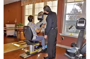 A person seated on a stationary exercise bike in a fitness or rehabilitation room, being assisted by another person standing next to them. The room has large windows with a view outside, wooden flooring, and exercise equipment visible.