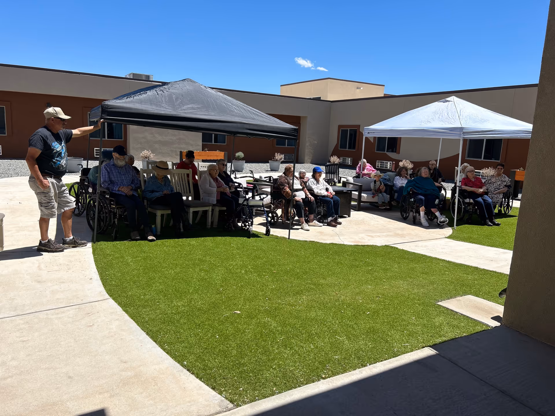 A group of elderly residents sitting under two canopy tents in a sunny assisted-living courtyard.