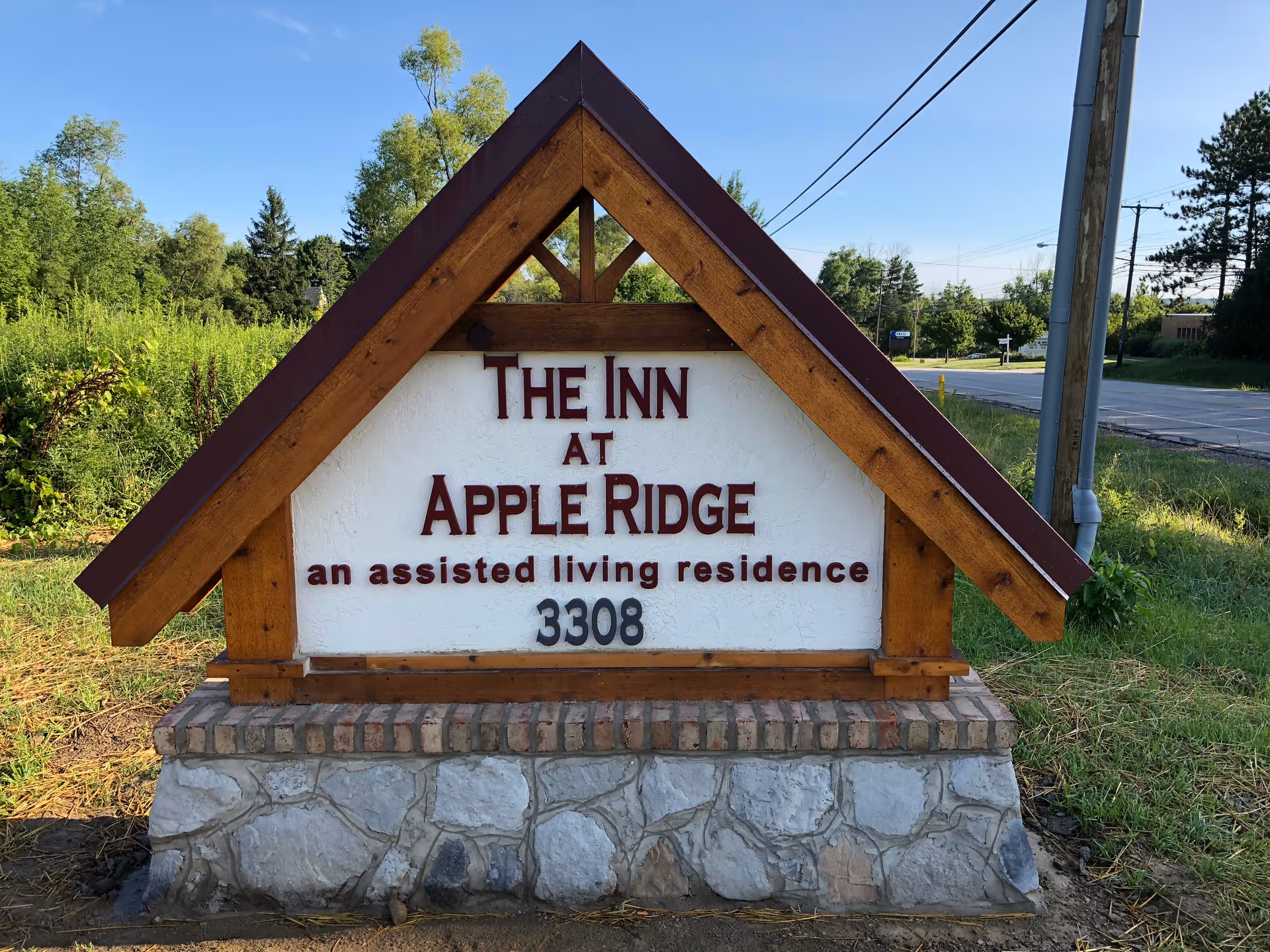 A wooden-framed roadside sign reading 'The Inn at Apple Ridge an assisted living residence 3308' with trees and a road in the background.