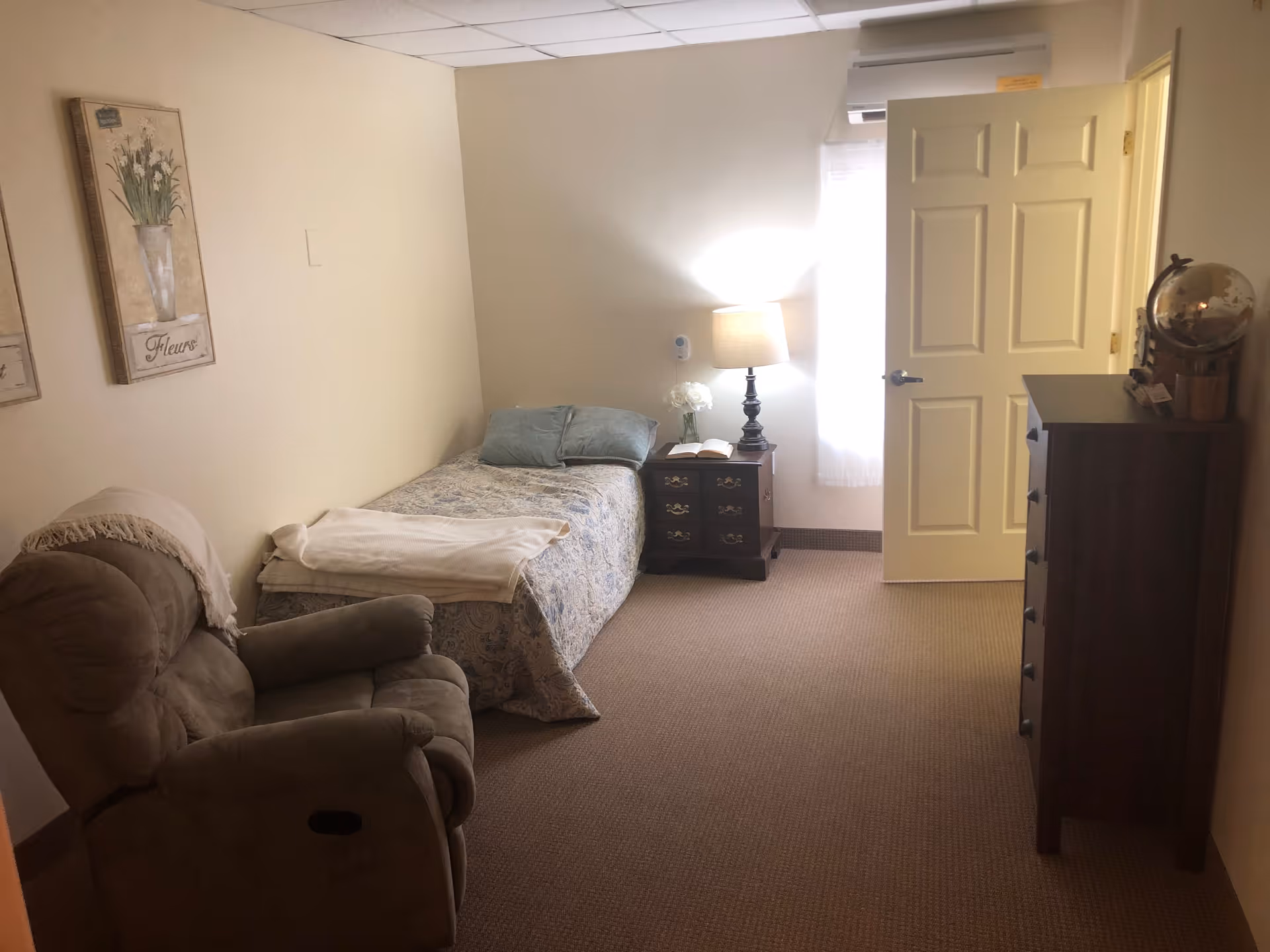 A small, cozy bedroom in a senior living community featuring a single bed with a patterned bedspread and two pillows, a wooden nightstand with a lamp, a book, and a vase with flowers. There is a comfortable recliner chair with a blanket draped over the back, a wooden dresser with a globe on top, and two framed floral artworks on the wall. The room has beige walls and carpeted floor, with a door partially open letting in natural light.