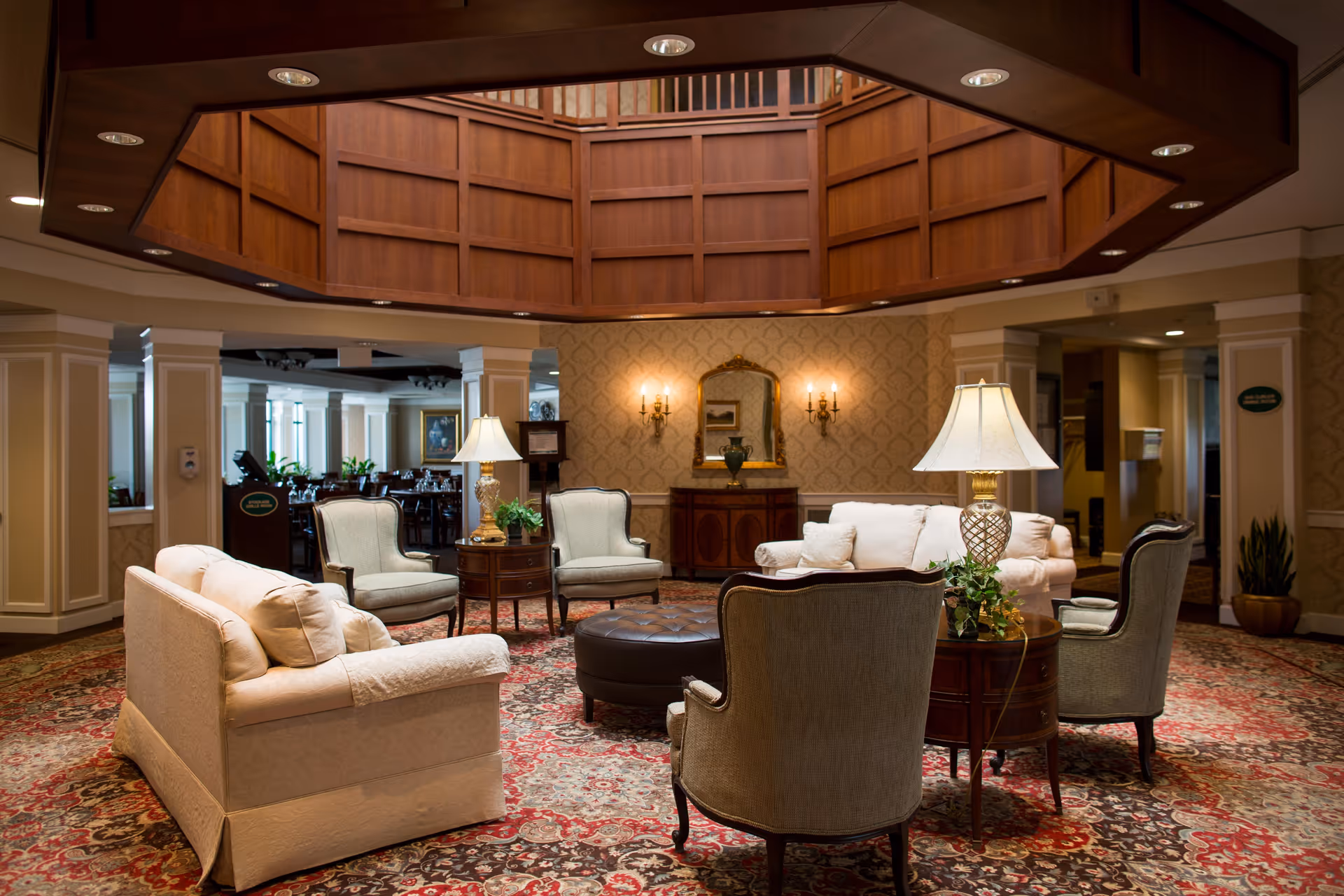 A cozy and elegant living room area with beige sofas and armchairs arranged around a round ottoman. Two table lamps with white shades sit on wooden side tables, and a patterned carpet covers the floor. The walls feature classic wallpaper, a mirror, and wall sconces with lights. The ceiling has a wooden architectural feature with recessed lighting.