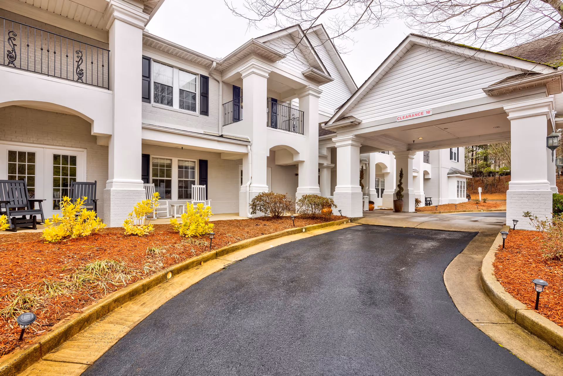 Exterior view of TerraBella Roswell facility showing a driveway leading to a covered entrance with white columns. The building has white walls, black shutters, and balconies. There are yellow flowering shrubs and mulch beds along the driveway.