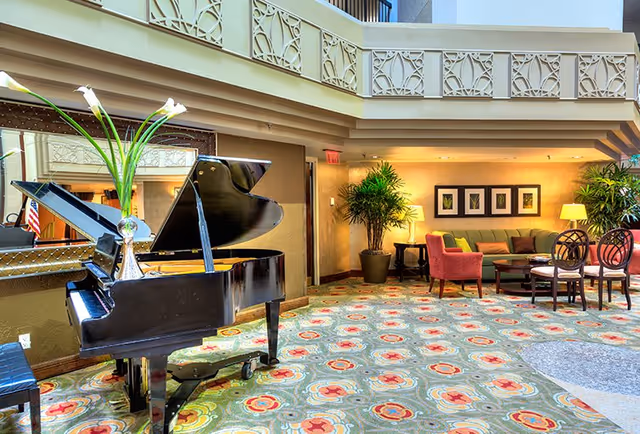 A bright and spacious living room area featuring a black grand piano with a vase of white flowers on top, colorful patterned carpet, a seating area with a green sofa, red armchairs, wooden chairs, coffee tables, potted plants, framed artwork on the wall, and warm lighting from lamps.