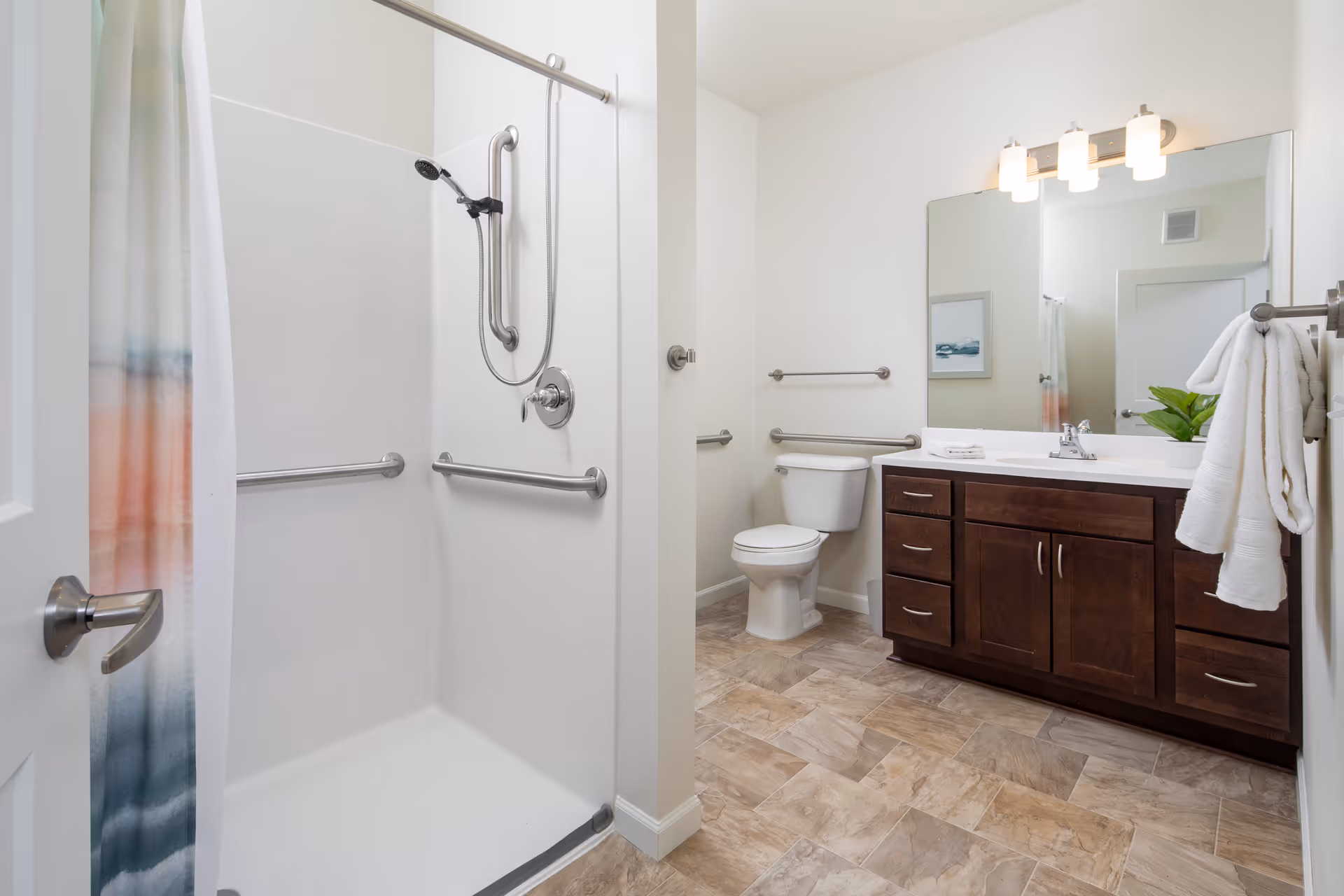 A clean and modern bathroom featuring a walk-in shower with grab bars and a handheld showerhead, a toilet with grab bars on the walls, and a wooden vanity with a large mirror and three-light fixture above. The floor has a tile pattern, and a towel is hanging on a rack next to the vanity.