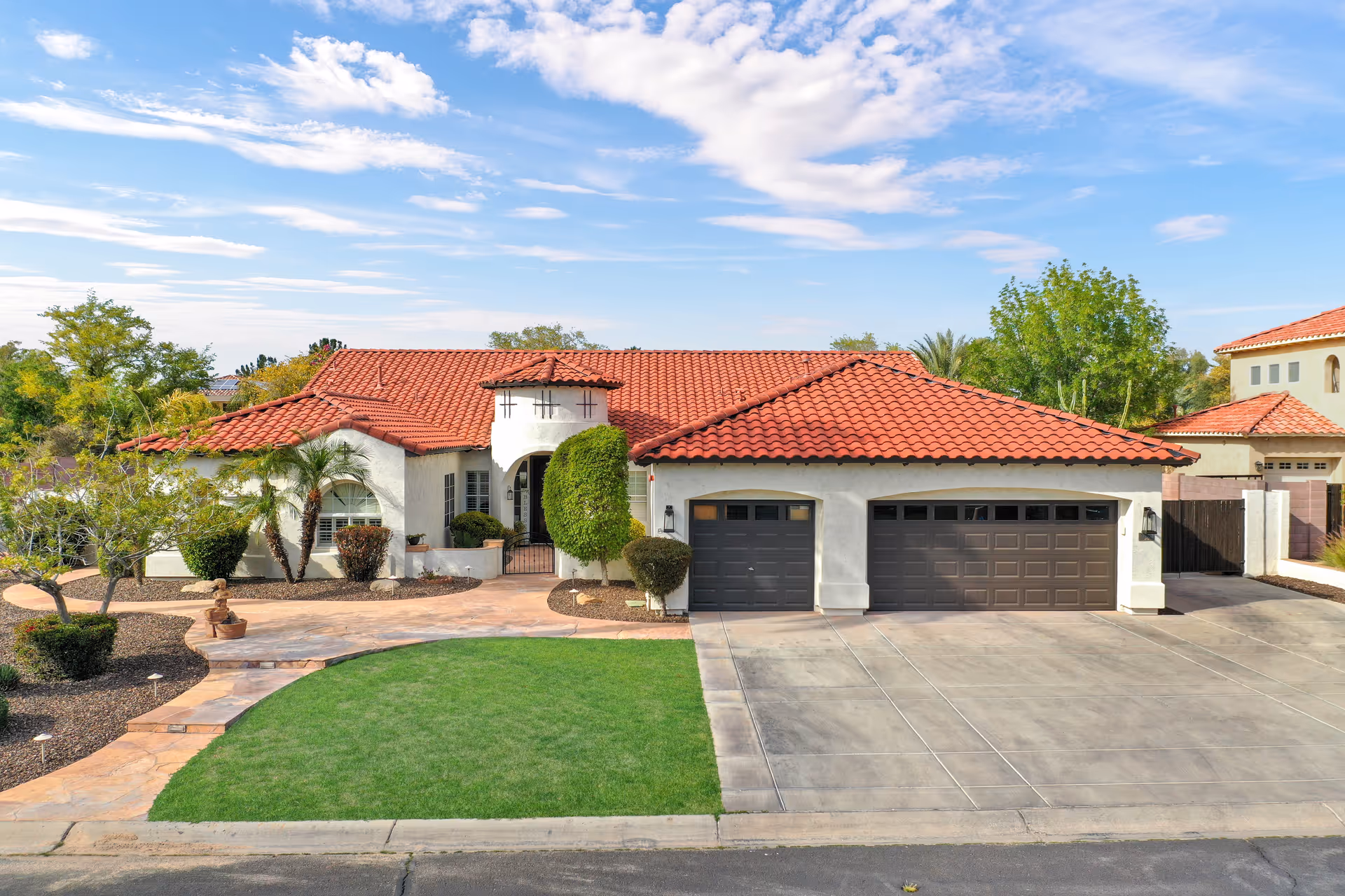 Front exterior view of a single-story senior living facility with a red tile roof, white walls, three dark garage doors, a well-maintained green lawn, and landscaped bushes and trees under a partly cloudy blue sky.