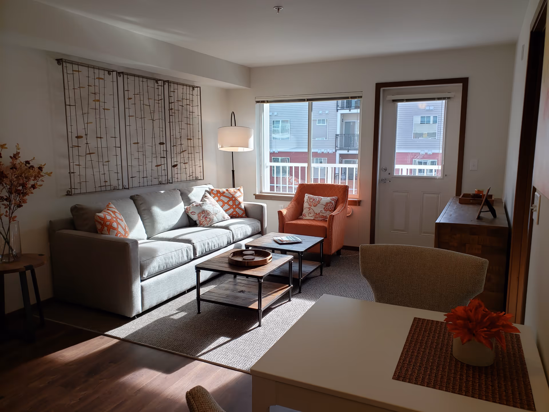 A cozy living room with a light gray sofa adorned with orange and patterned pillows, an orange armchair with a decorative pillow, two wooden coffee tables, a floor lamp, and a window and door leading to a balcony. The room features a wooden sideboard, a small table with a red flower centerpiece, and wall art with a nature-inspired design.