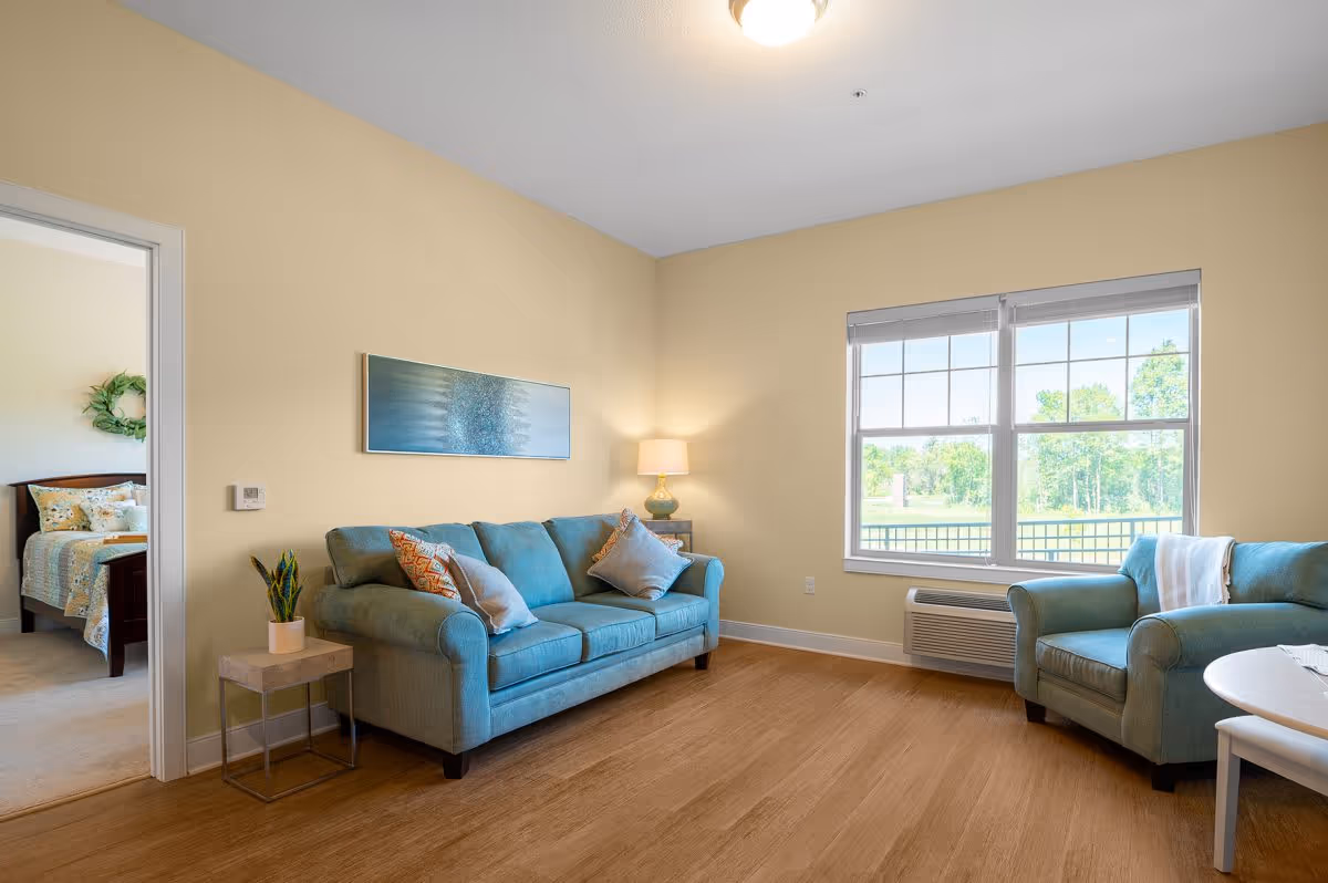 Sunlit living room with teal sofa and armchair, wooden floor, lamp, and a large window overlooking greenery.