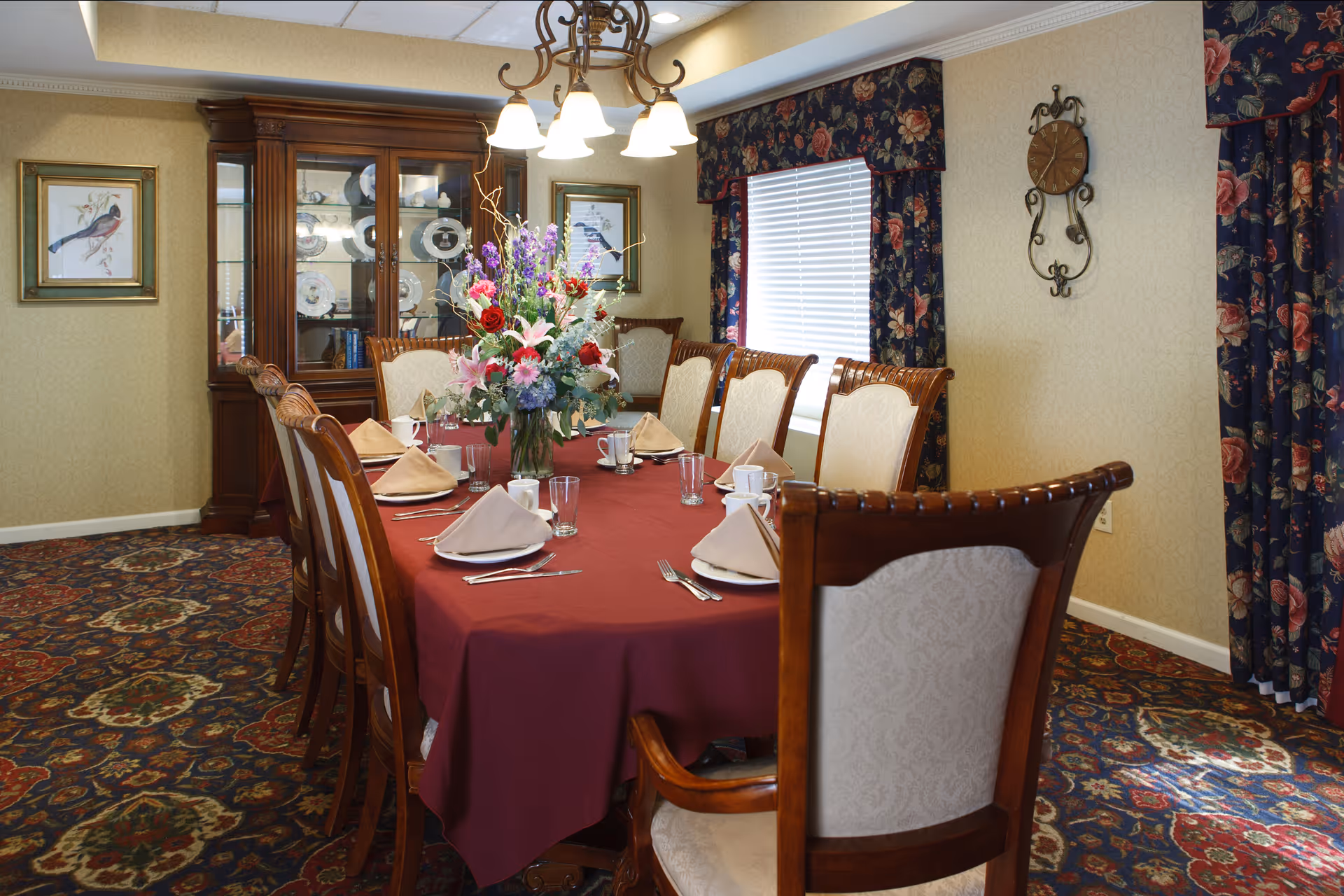 A formal dining room with a long table set with a burgundy tablecloth, napkins, place settings and a large floral centerpiece, surrounded by wooden chairs and a china cabinet.
