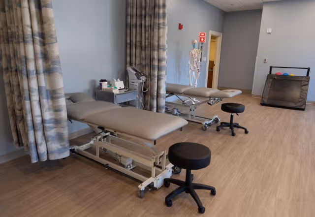 A medical treatment room with two adjustable examination tables separated by patterned curtains. Each table has a black rolling stool nearby. Medical equipment and supplies are placed on a small cart next to one of the tables. A human skeleton model is visible near the back wall, along with a fire alarm and an open doorway. The floor is wood-patterned, and the walls are painted light blue.