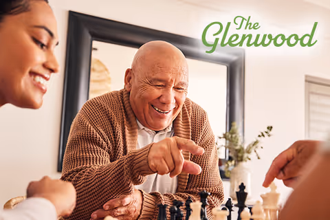 An elderly man wearing a brown cardigan smiling and playing chess with two other people in a bright room with a large mirror and a plant in the background.