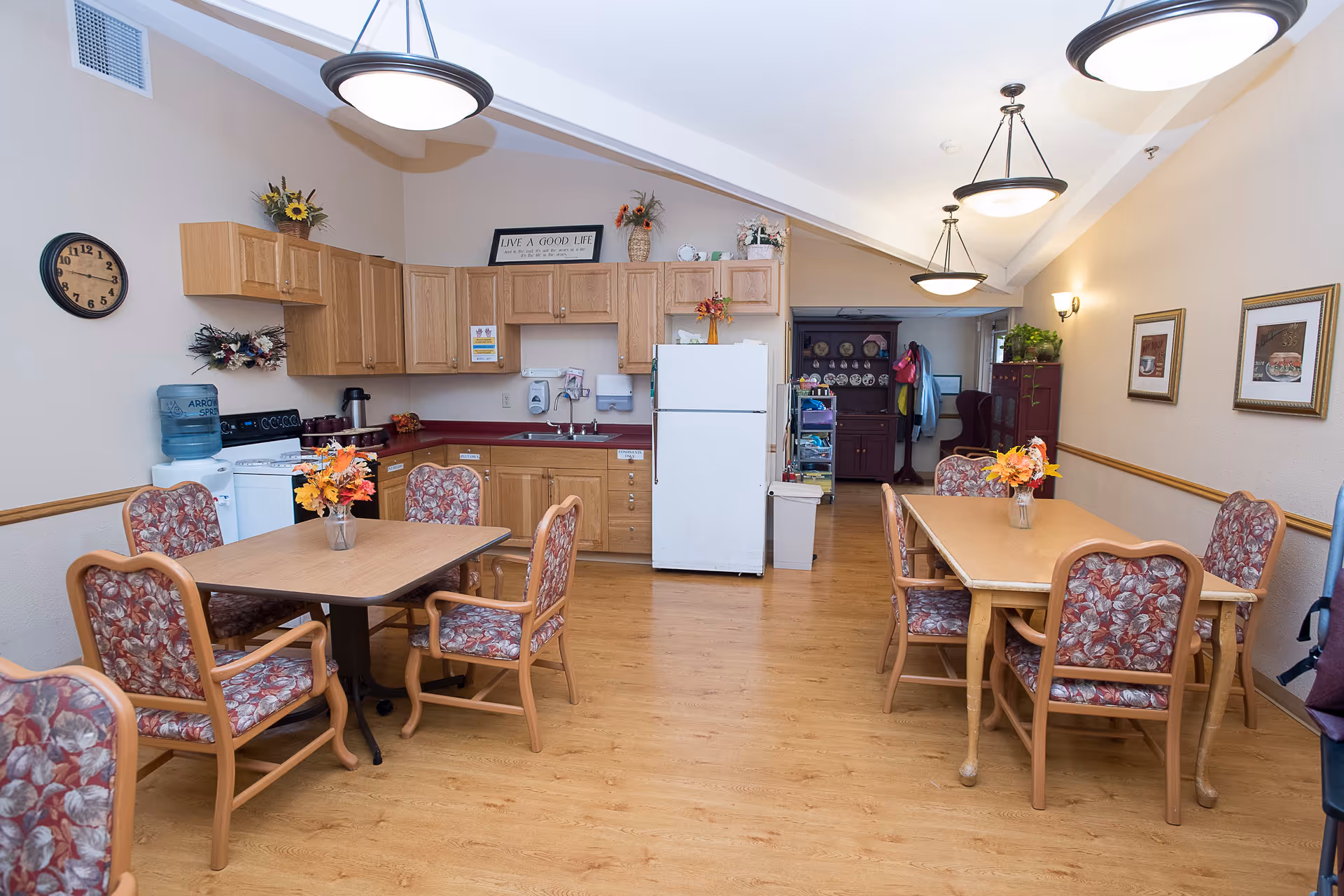 A cozy kitchen and dining area in a senior living facility with wooden cabinets, a white refrigerator, a stove, and a water dispenser. There are two dining tables with floral upholstered chairs and flower arrangements on each table. The room has wooden flooring, beige walls with framed pictures, and ceiling lights.