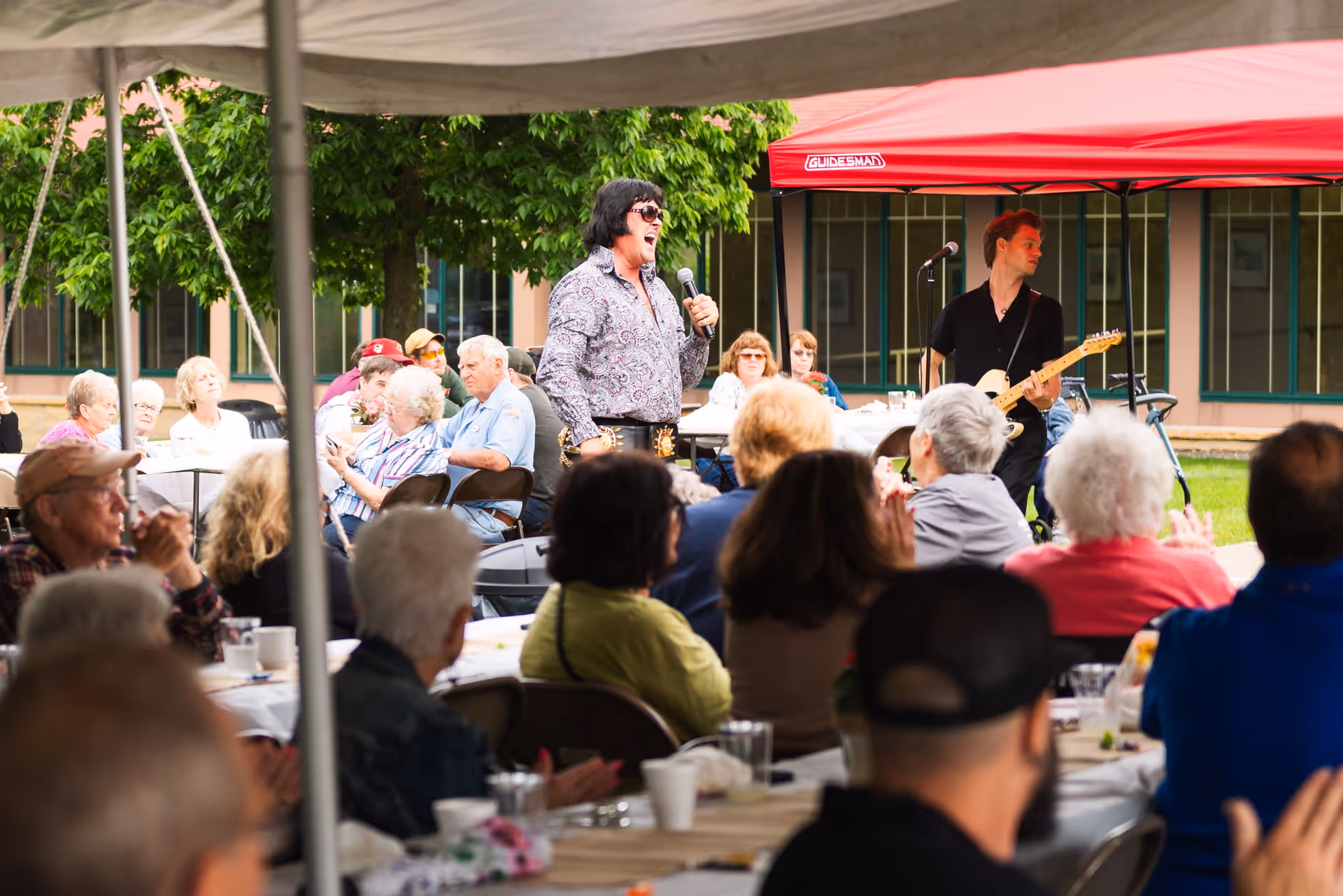 A group of elderly people seated at tables outdoors under a tent, watching a live musical performance. A man dressed like Elvis Presley is singing into a microphone, and another man is playing an electric guitar under a red canopy. The setting appears to be a social event at a senior living facility with greenery and building windows in the background.