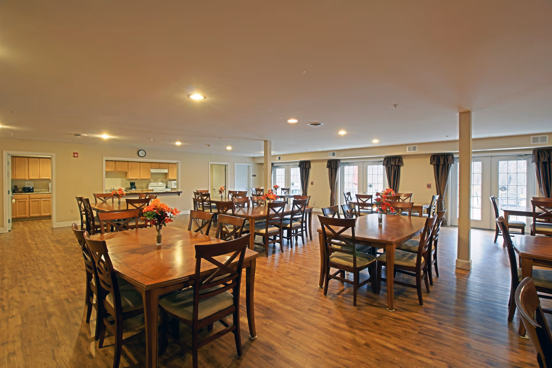 Spacious dining room with multiple wooden tables and chairs arranged neatly. Each table has a small vase with orange flowers. The room has wood flooring, beige walls, and several large windows with dark curtains allowing natural light to enter. A kitchen area is visible through a serving window on one side of the room.