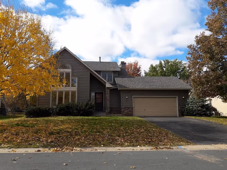 A two-story suburban house with a large garage, surrounded by trees with autumn foliage and a lawn covered with fallen leaves under a partly cloudy sky.