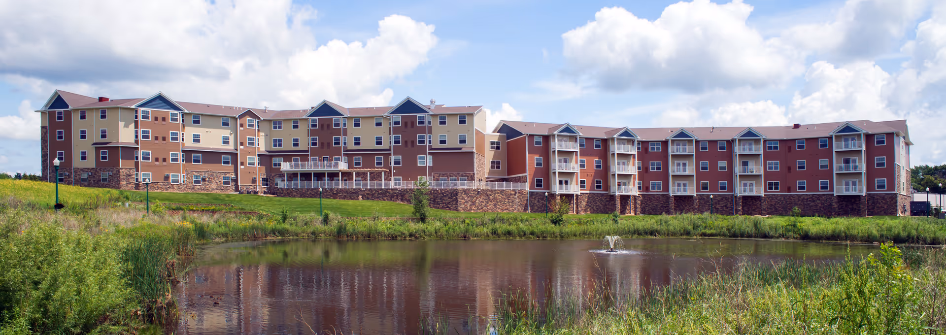 Wide exterior view of a large multi-story senior living facility building with beige, red, and brown colors, situated behind a pond with a small fountain, surrounded by green grass and vegetation under a partly cloudy sky.