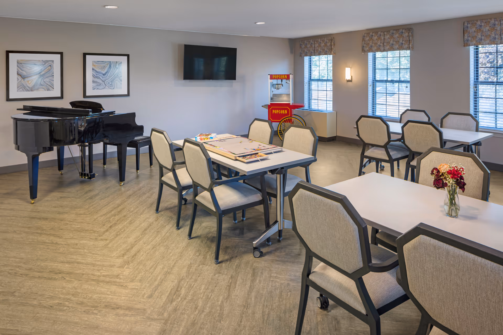 Bright communal activity/dining room with tables and chairs, a grand piano, TV, and a popcorn machine.
