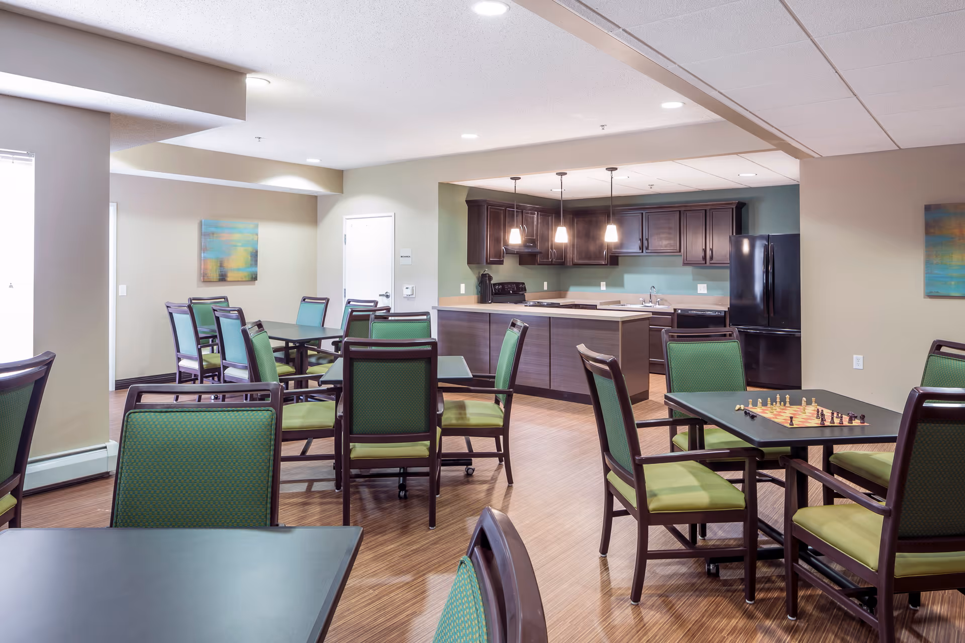 A communal dining and kitchen area in a senior living facility with multiple tables and green cushioned chairs. The kitchen features dark wood cabinets, a black refrigerator, a stove, and a dishwasher. One table has a chessboard set up. The room has wood flooring, beige walls, and modern ceiling lights.