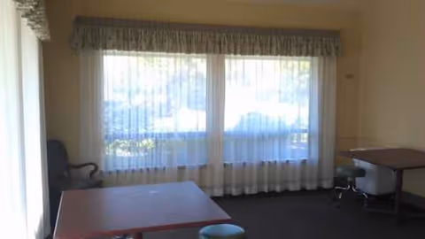 Sunlit senior living common area with large windows, sheer curtains, and tables.