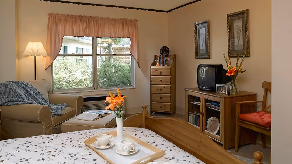 A cozy senior living room with a beige armchair draped with a blue blanket, a floor lamp, a window with pink curtains, a wooden dresser and TV stand with a small TV, framed pictures on the wall, a wooden chair with a red cushion, and a bed with a tray holding two teacups and a vase with orange flowers.