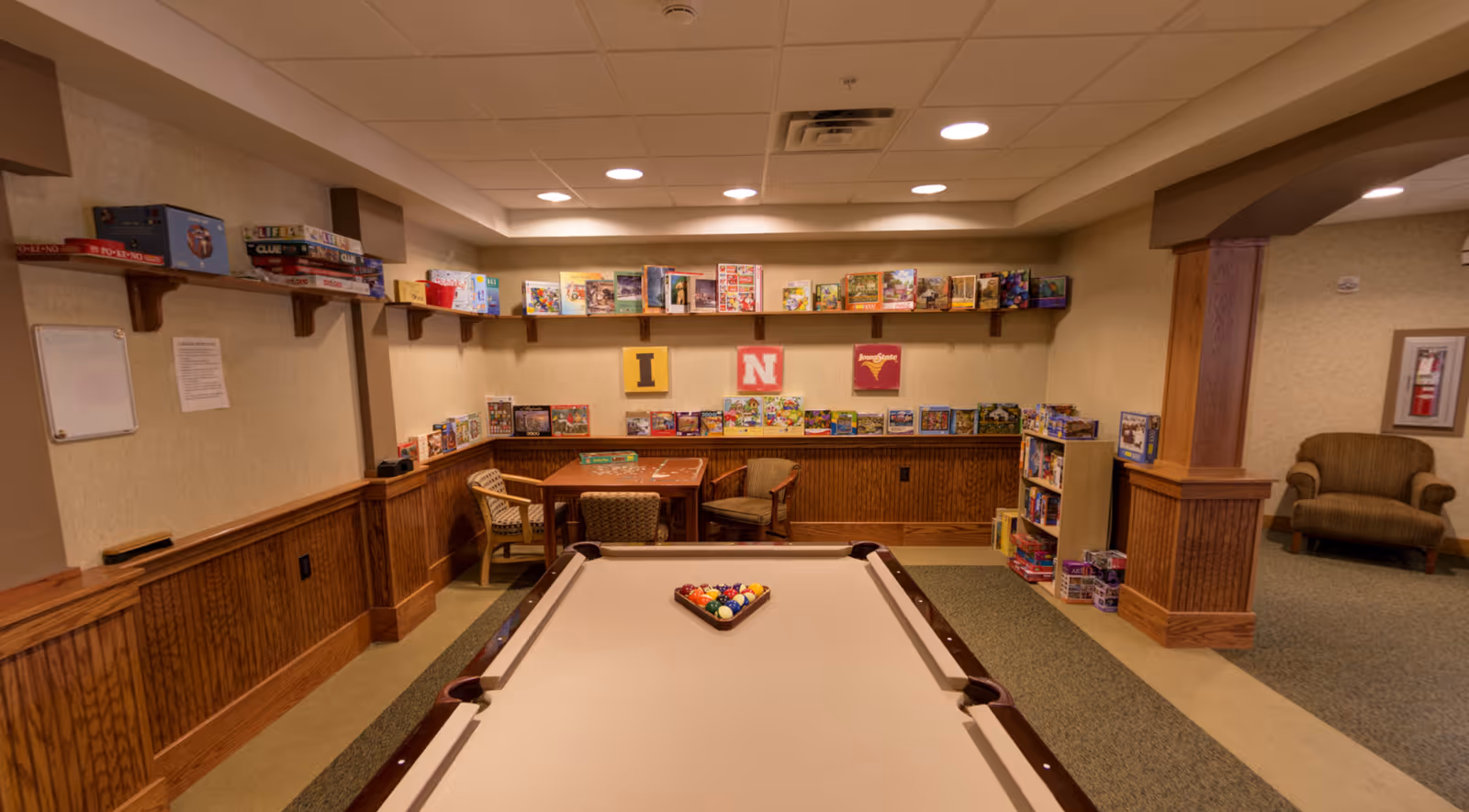 A recreational room with a pool table in the foreground, a table with chairs, and shelves filled with board games and puzzles along the walls. The room has wood paneling on the lower walls and beige wallpaper above, with recessed ceiling lights providing illumination.