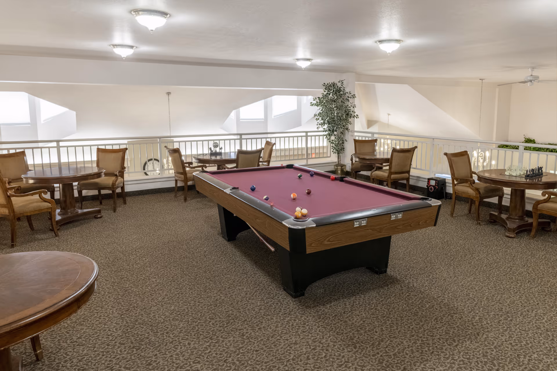 A senior living community game room featuring a pool table with a purple felt top in the center. Surrounding the pool table are several round wooden tables with upholstered chairs. The room has beige walls, carpeted flooring, ceiling lights, and a potted plant near the railing overlooking a lower level.