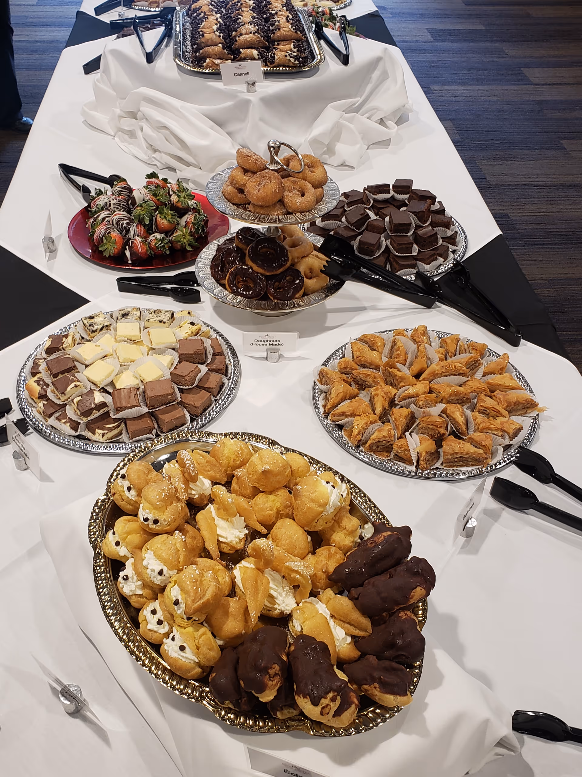 A buffet table with a white tablecloth displaying an assortment of desserts including chocolate-covered strawberries, mini donuts, brownies, cannoli, cream puffs, and other pastries arranged on silver platters with serving tongs.