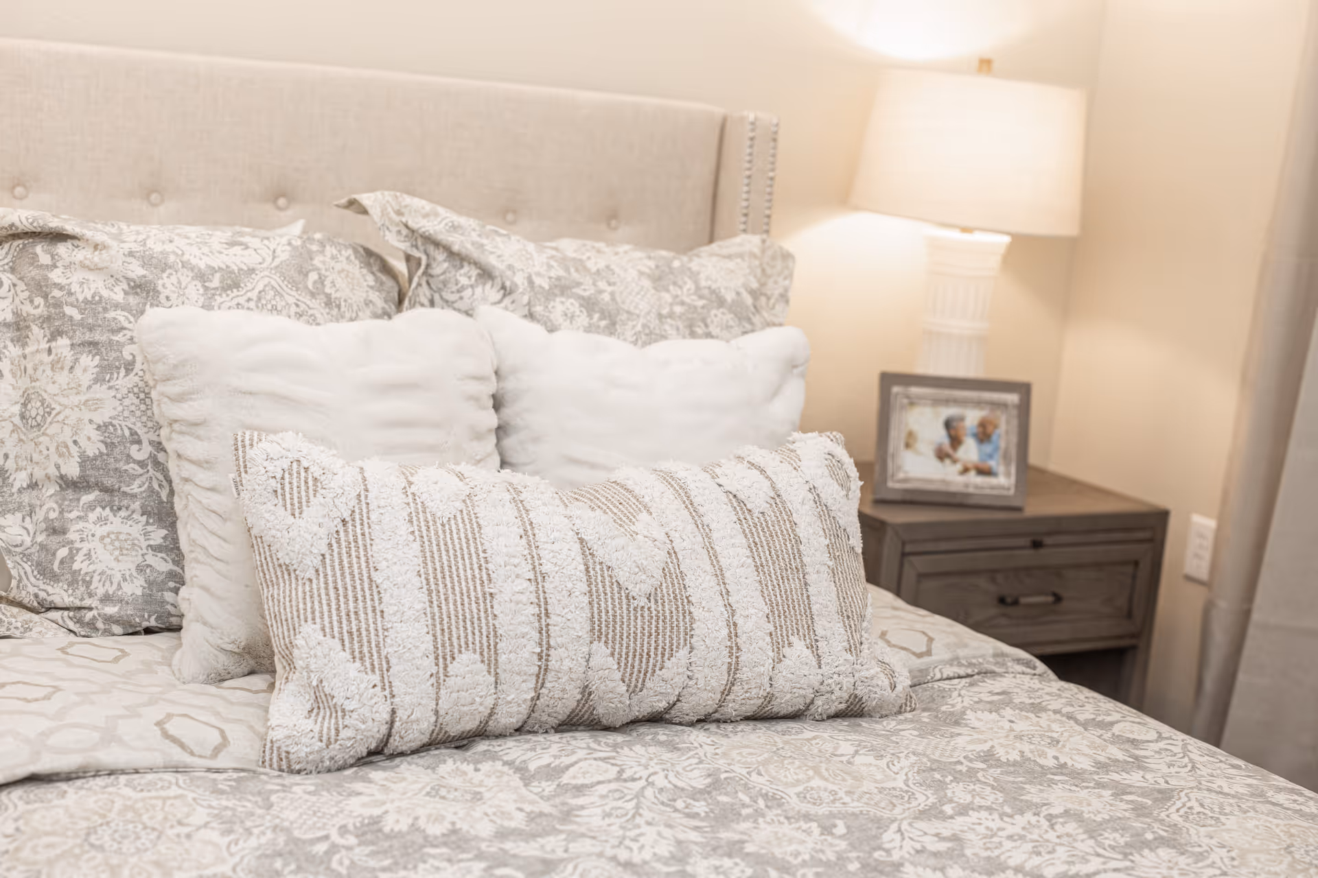 A neatly made bed with multiple decorative pillows in shades of white and beige, a beige upholstered headboard, and a wooden nightstand with a lamp and a framed photo in the background.