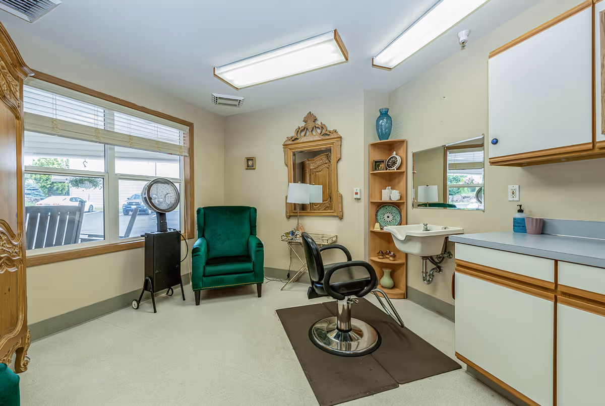 A small salon room with a black salon chair on a mat in front of a wall-mounted sink and mirror. To the left, there is a green upholstered armchair next to a vintage hair dryer. The room has light-colored walls, a large window with blinds, wooden cabinetry, and decorative items on shelves including a blue vase and plates.