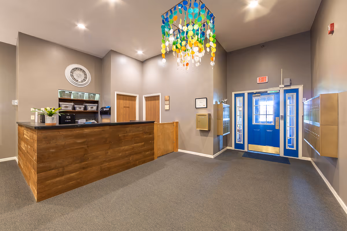 Reception area of a senior living facility with a wooden front desk, a decorative clock on the wall, and a colorful hanging light fixture. There are two wooden doors behind the desk, a blue entrance door with glass panels, mailboxes on the right wall, and a gray carpeted floor.