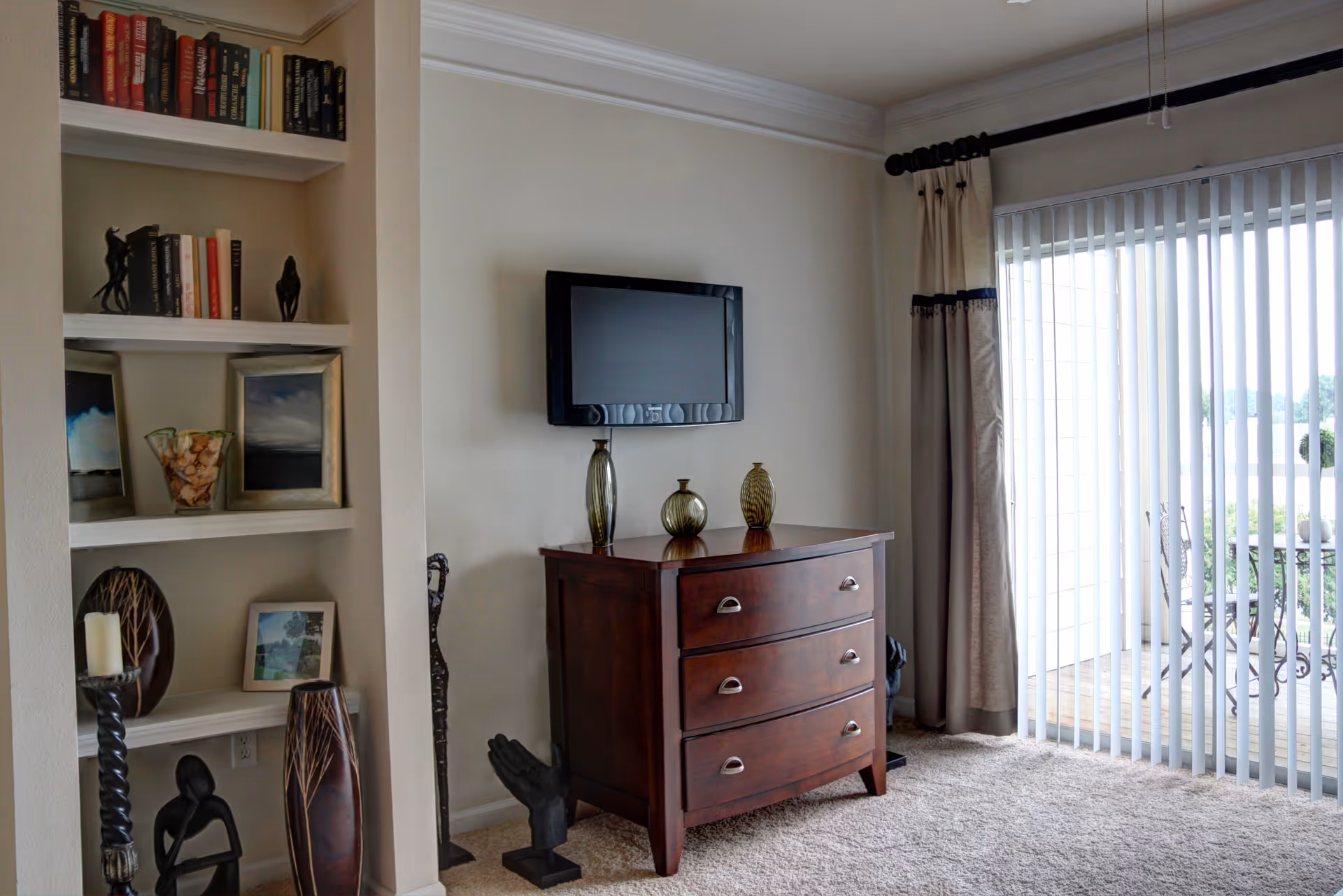 A cozy living room area featuring a wooden chest of drawers with three glass decorative vases on top, a wall-mounted flat-screen TV above it, built-in shelves filled with books, framed pictures, and decorative items on the left, and a large sliding glass door with vertical blinds leading to an outdoor patio with a table and chairs.