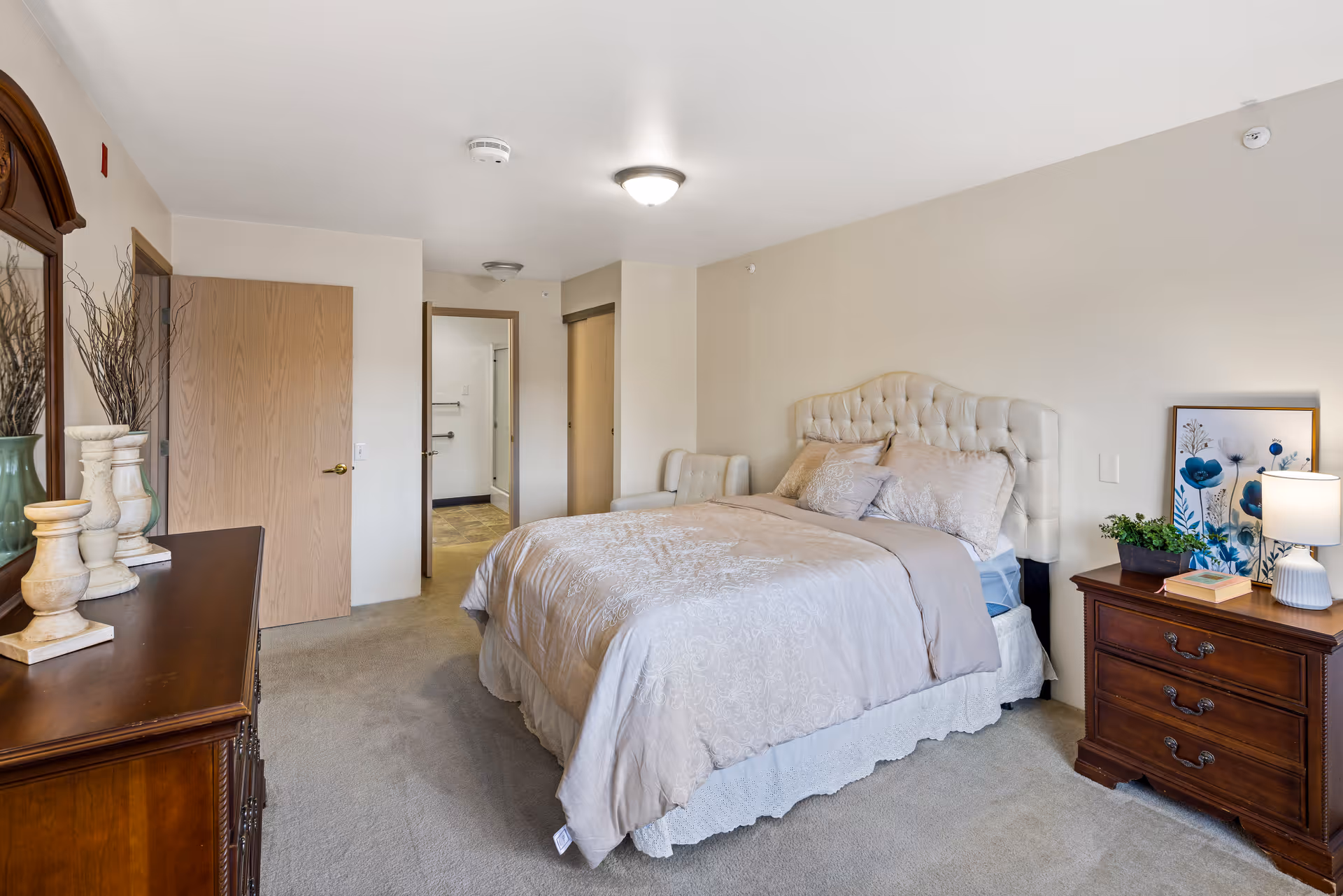 A neatly arranged bedroom in a senior living community featuring a large bed with a tufted headboard, beige bedding, and multiple pillows. To the right of the bed is a wooden nightstand with a lamp, a small plant, a book, and framed floral artwork. On the left side of the room is a wooden dresser with decorative vases and a mirror above it. The room has beige walls, carpeted floor, and two open doors leading to other areas of the facility.