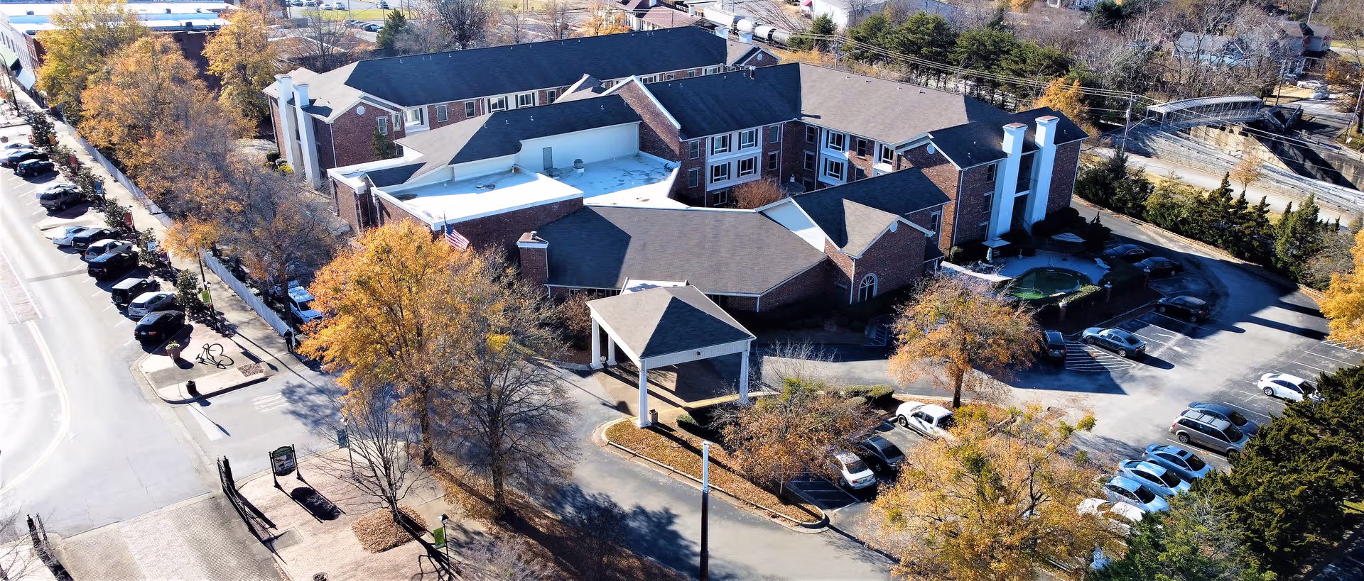Aerial view of City Center Village Senior Community, showing a large multi-wing brick building with a covered entrance, surrounded by trees with autumn foliage and parking lots filled with cars.