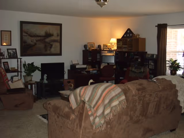 A cozy living room with brown upholstered sofas and armchairs, a striped throw blanket draped over the back of one sofa, a small TV on a stand, a wooden shelving unit with framed photos and plants, a desk with office supplies and a lamp, and a window with dark curtains letting in natural light.