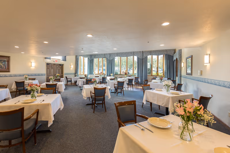 Spacious dining room with multiple tables covered in white linens and floral centerpieces, chairs, and large windows along the back wall.