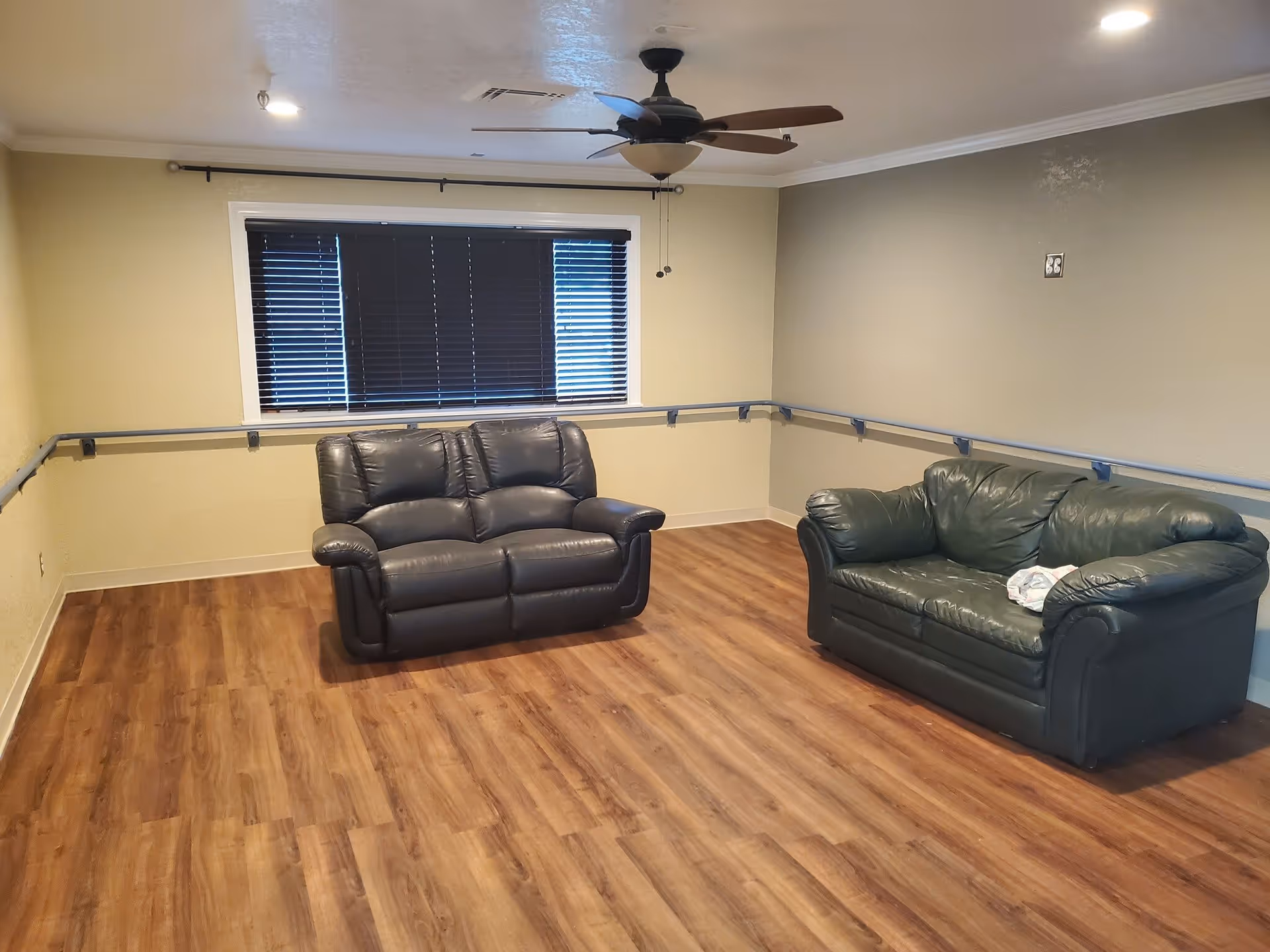 A room with two leather sofas, one black and one dark green, placed on a wooden floor. The walls are painted beige and gray, with a window covered by dark blinds. A ceiling fan with lights is mounted on the ceiling, and there are handrails along the walls.