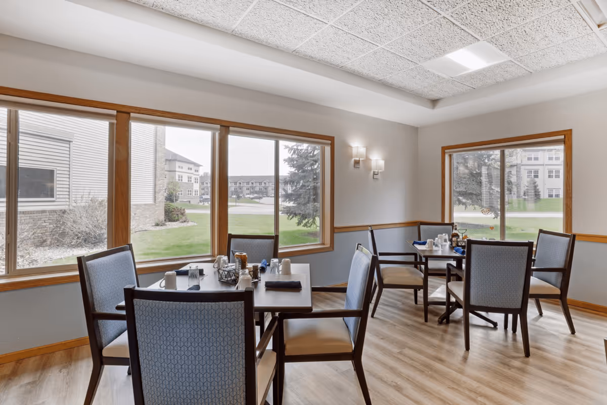 A bright dining room in a senior living facility with two tables, each surrounded by four chairs. The tables are set with cups, napkins, and condiments. Large windows provide a view of the green lawn and neighboring buildings outside. The room has light-colored walls, wooden trim, and a light wood floor.