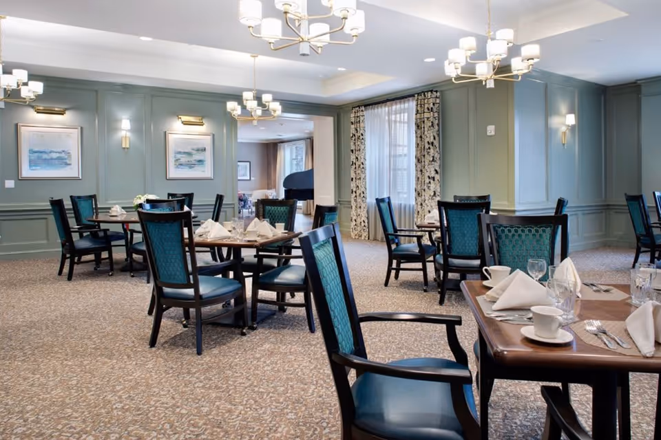 A well-lit dining room with multiple round and square tables set with white napkins, cups, glasses, and silverware. The room features green paneled walls, patterned carpet, framed artwork, and large windows with floral curtains. Elegant chandeliers and wall sconces provide lighting, and a grand piano is visible in an adjoining room.