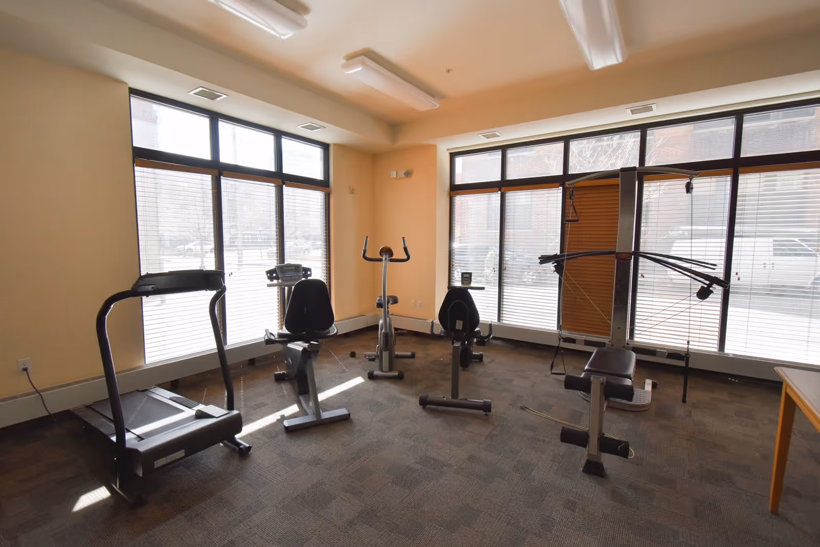 A small fitness room with large windows letting in natural light. The room contains a treadmill, two stationary bikes, and a multi-function weight machine. The walls are painted beige and the floor is carpeted.