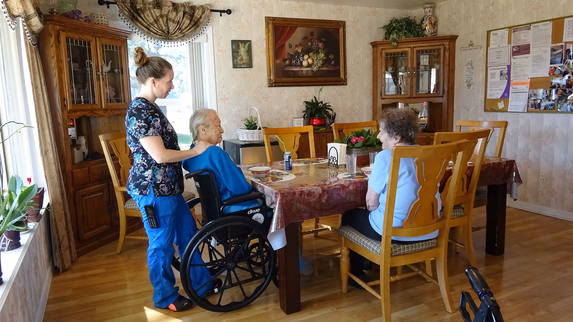 A caregiver standing behind an elderly man in a wheelchair at a dining table, with an elderly woman sitting across from them. The room has wooden furniture, a large window with curtains, a cabinet with glass doors, and a bulletin board on the wall.