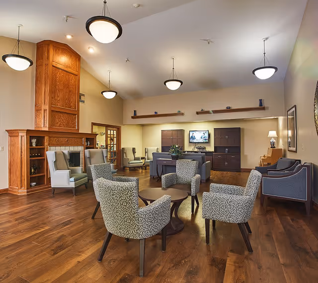 A spacious senior living facility common area with wooden flooring, multiple armchairs arranged around small tables, a large wooden fireplace on the left, and a TV mounted on the far wall above cabinets. The room is well-lit with hanging ceiling lights and has a warm, inviting atmosphere.