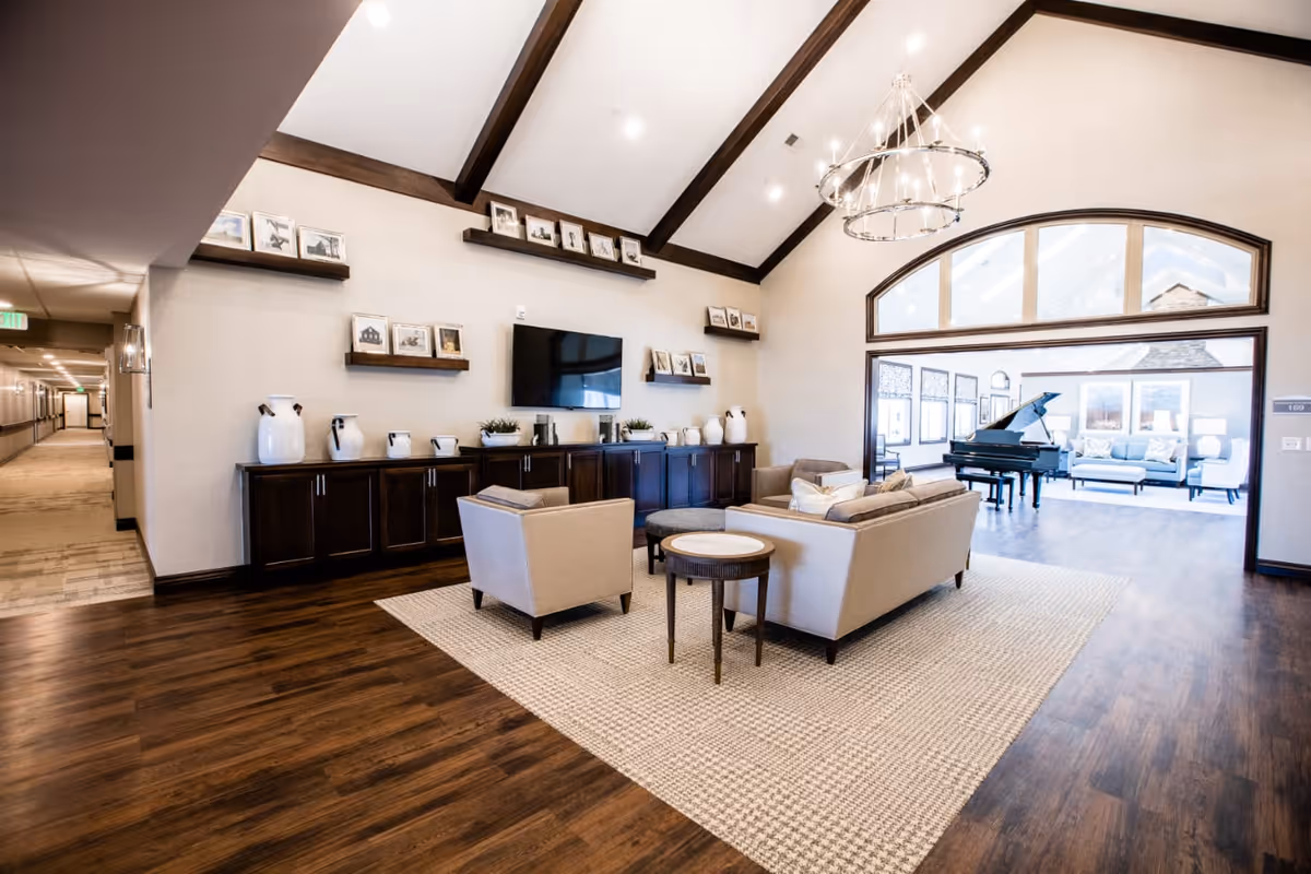A spacious senior living facility common area with high vaulted ceilings and exposed wooden beams. The room features a seating arrangement with beige sofas and armchairs on a large textured rug, a dark wooden cabinet adorned with white vases and framed photos, and a flat-screen TV mounted on the wall. In the background, through a large arched window, there is another room with a grand piano and additional seating. The flooring is dark wood, and a modern chandelier hangs from the ceiling.