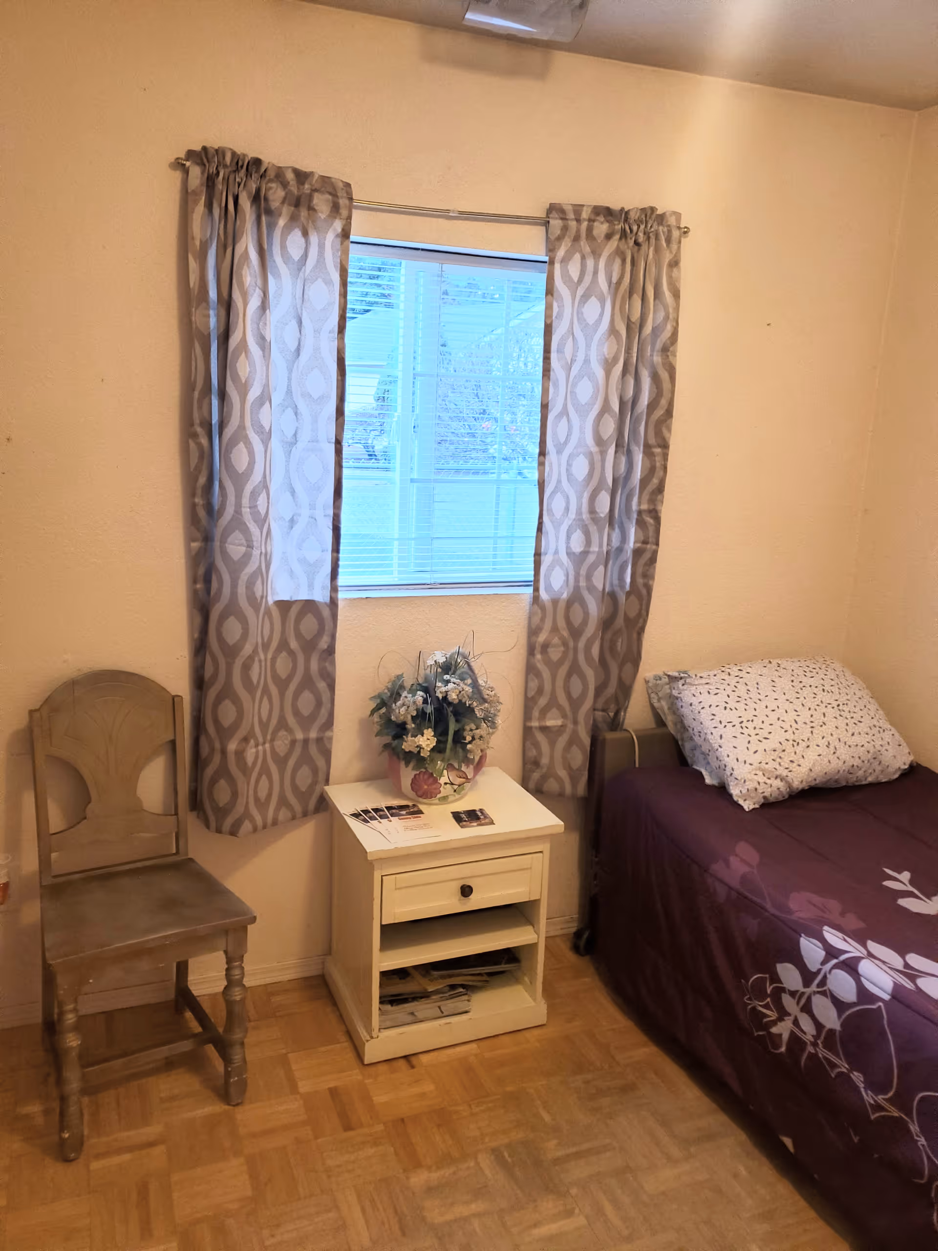 A small bedroom with a single bed covered in a purple floral bedspread and a white pillow with small patterns. Next to the bed is a white nightstand with a drawer and an open shelf containing some papers. On top of the nightstand is a vase with artificial flowers and some brochures. A wooden chair is placed to the left of the nightstand. The room has a window with patterned curtains partially drawn, allowing light to enter. The floor is wooden parquet.