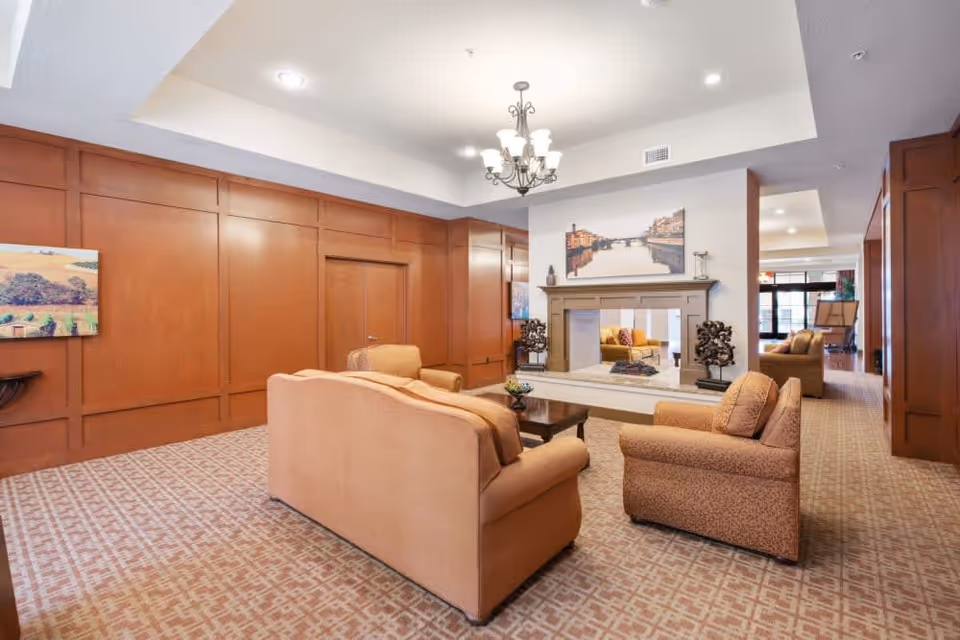 A cozy living room area with beige upholstered sofas and armchairs arranged around a wooden coffee table. The room features a decorative fireplace with a large framed picture above it, wood-paneled walls, a patterned carpet, and a chandelier hanging from the ceiling. There is additional seating visible in the background near large windows.